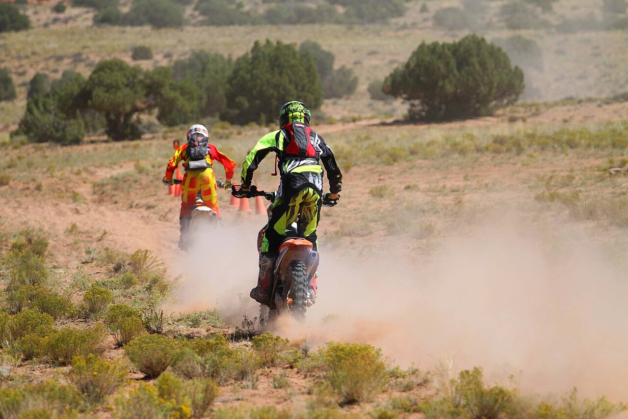 Two dirt bike riders kicking up dust on a desert trail among juniper trees