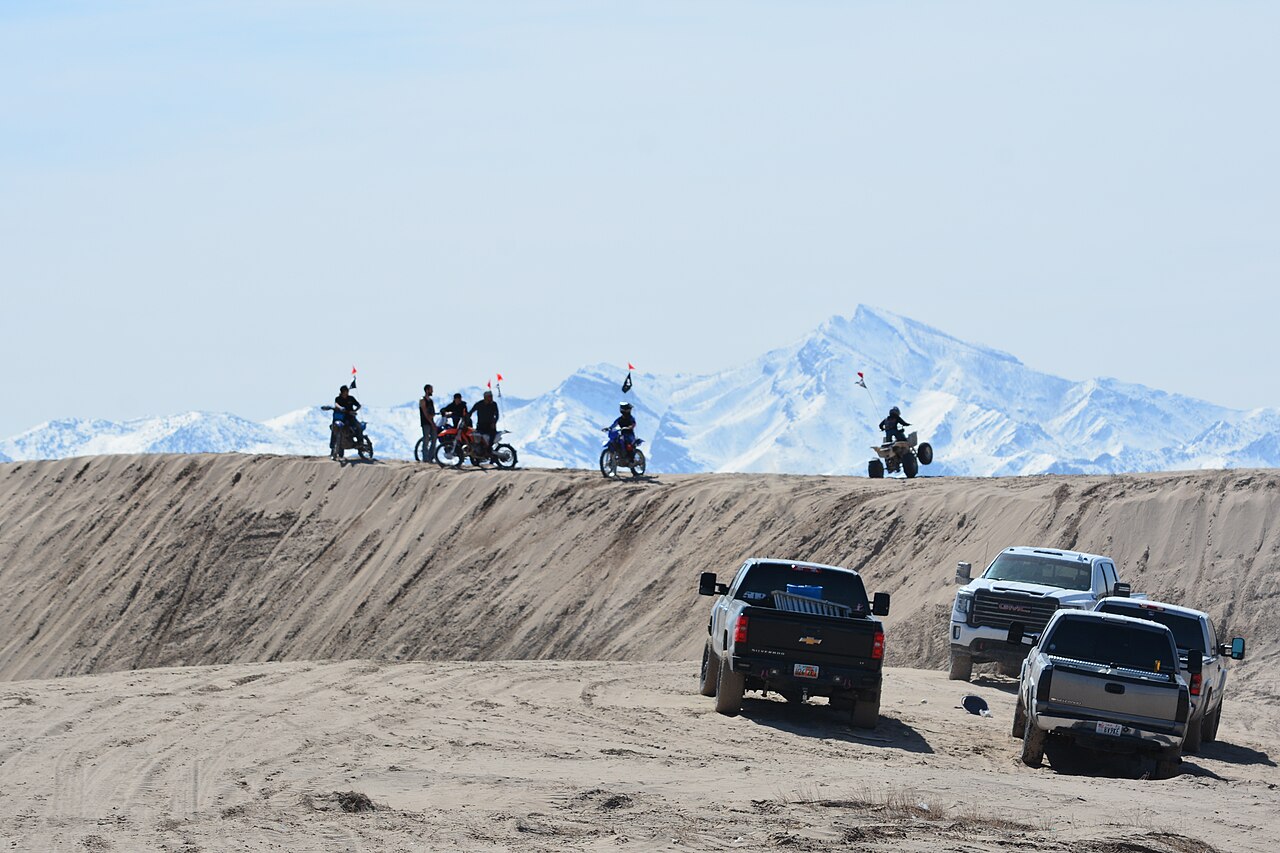 Dirt bike riders and an ATV lined up on a sand ridge at Little Sahara Recreation Area in Utah, with snowy mountains behind